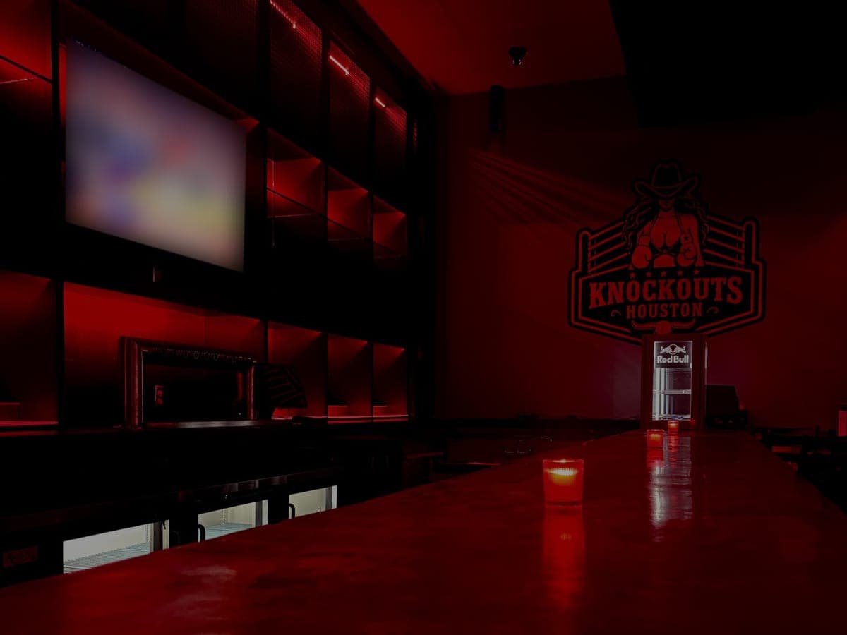 The long bar top at Knockouts Houston lit red, with candles along it, a TV above the back bar and the club's logo on the far wall
