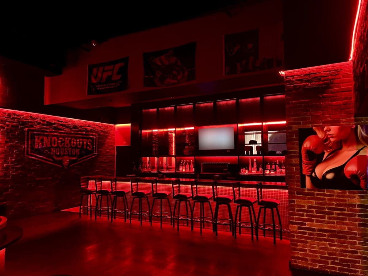 The bar at Knockouts Houston: a row of stools along a red-lit tiled bar, with the club's sign on a brick wall and sports banners above