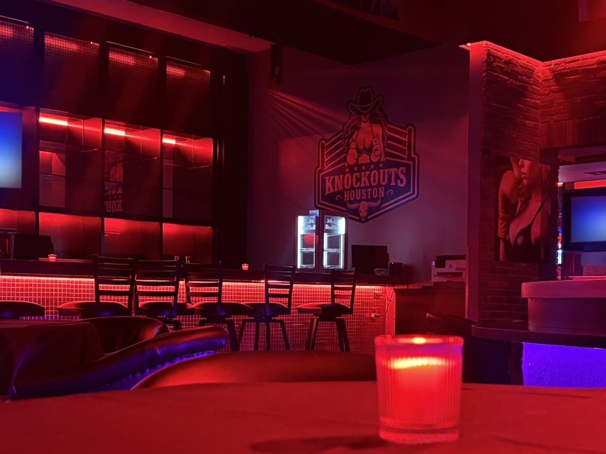 A candle on a table at Knockouts Houston, looking across the room to the bar stools and the club's logo on the wall